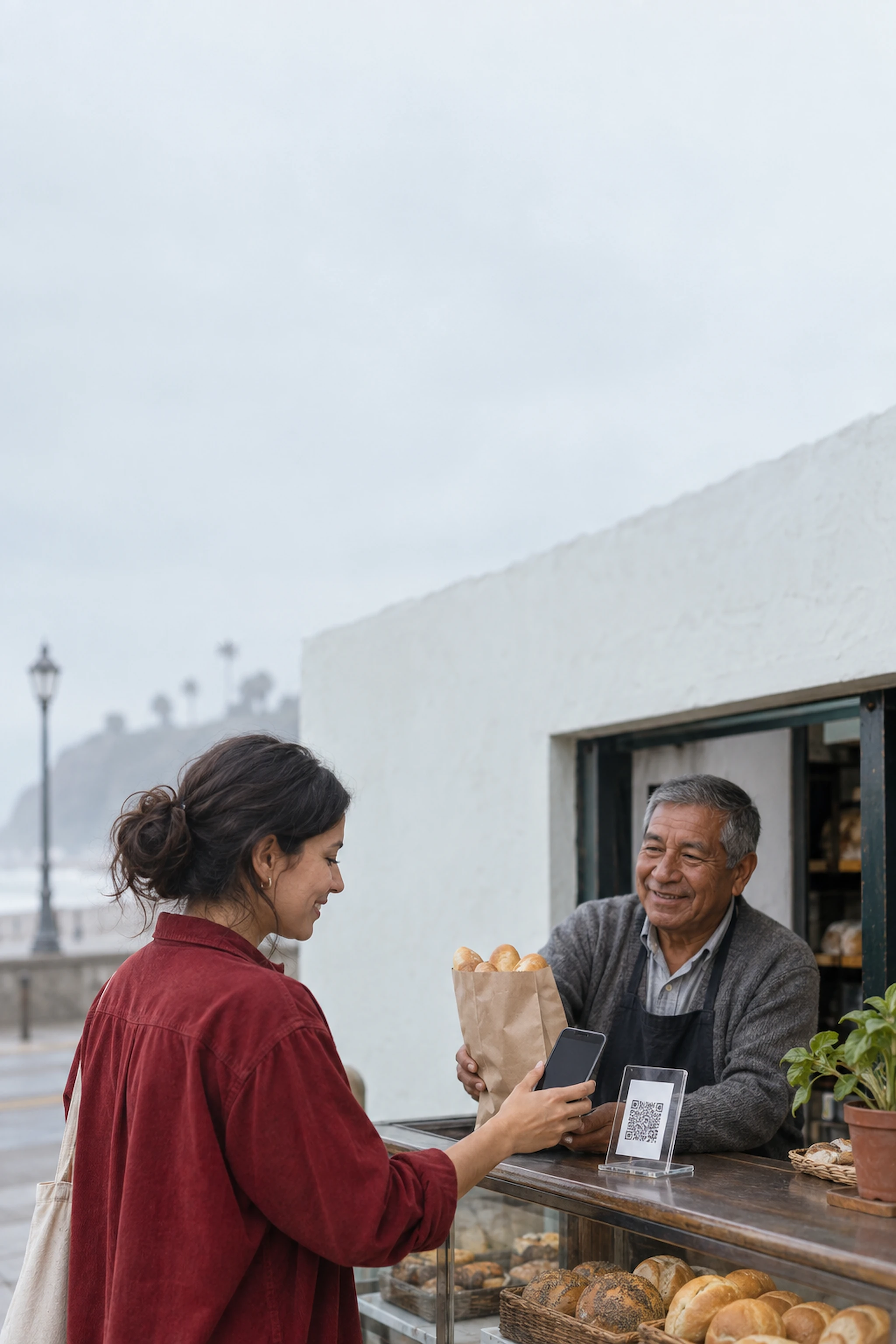A customer pays with her phone at a neighborhood bodega in Lima while the shopkeeper hands her a bag of bread.